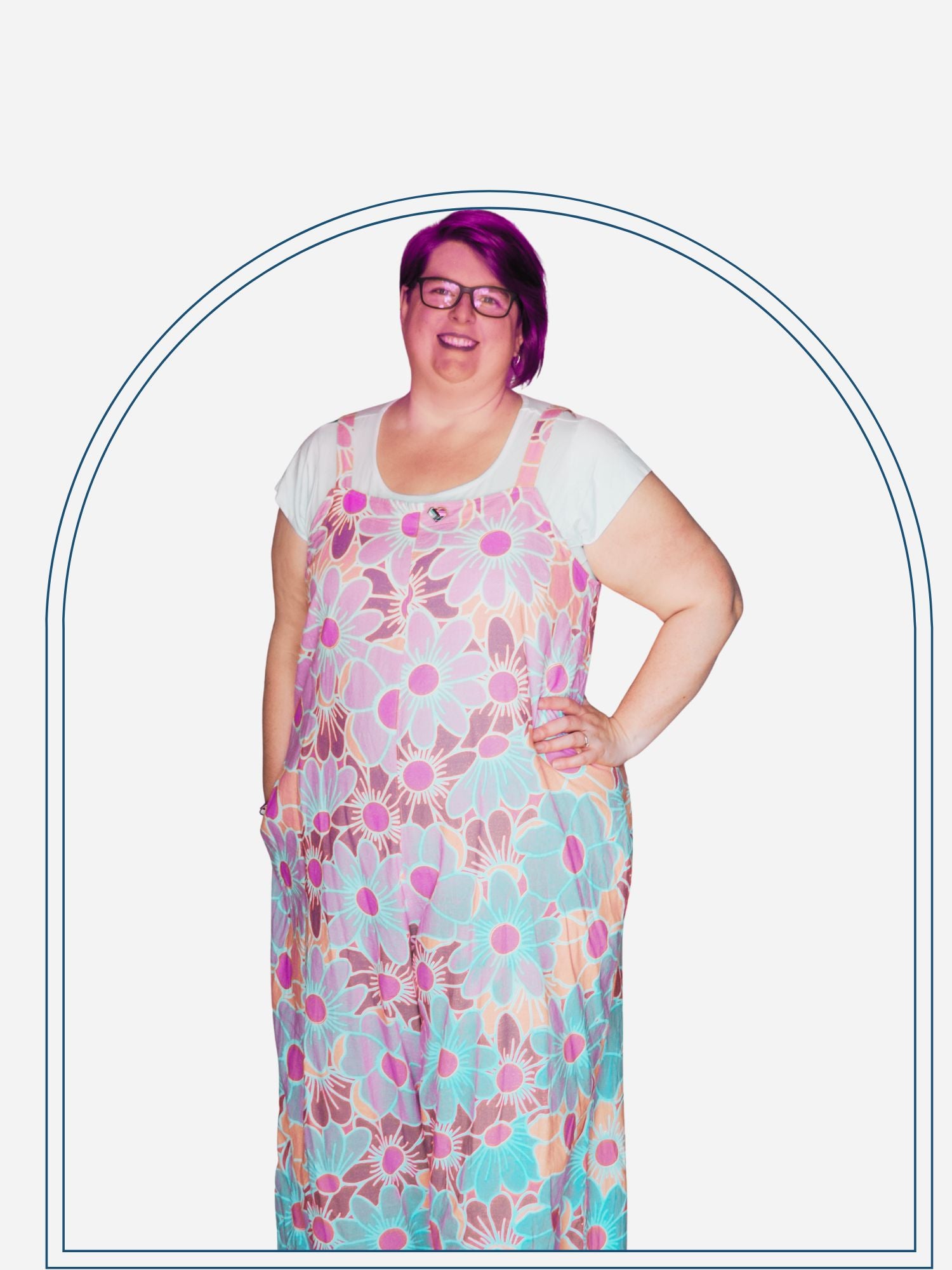 A woman standing in a doorway wearing a square neck jumpsuit with a colorful floral pattern on a purple background, paired with white sandals.