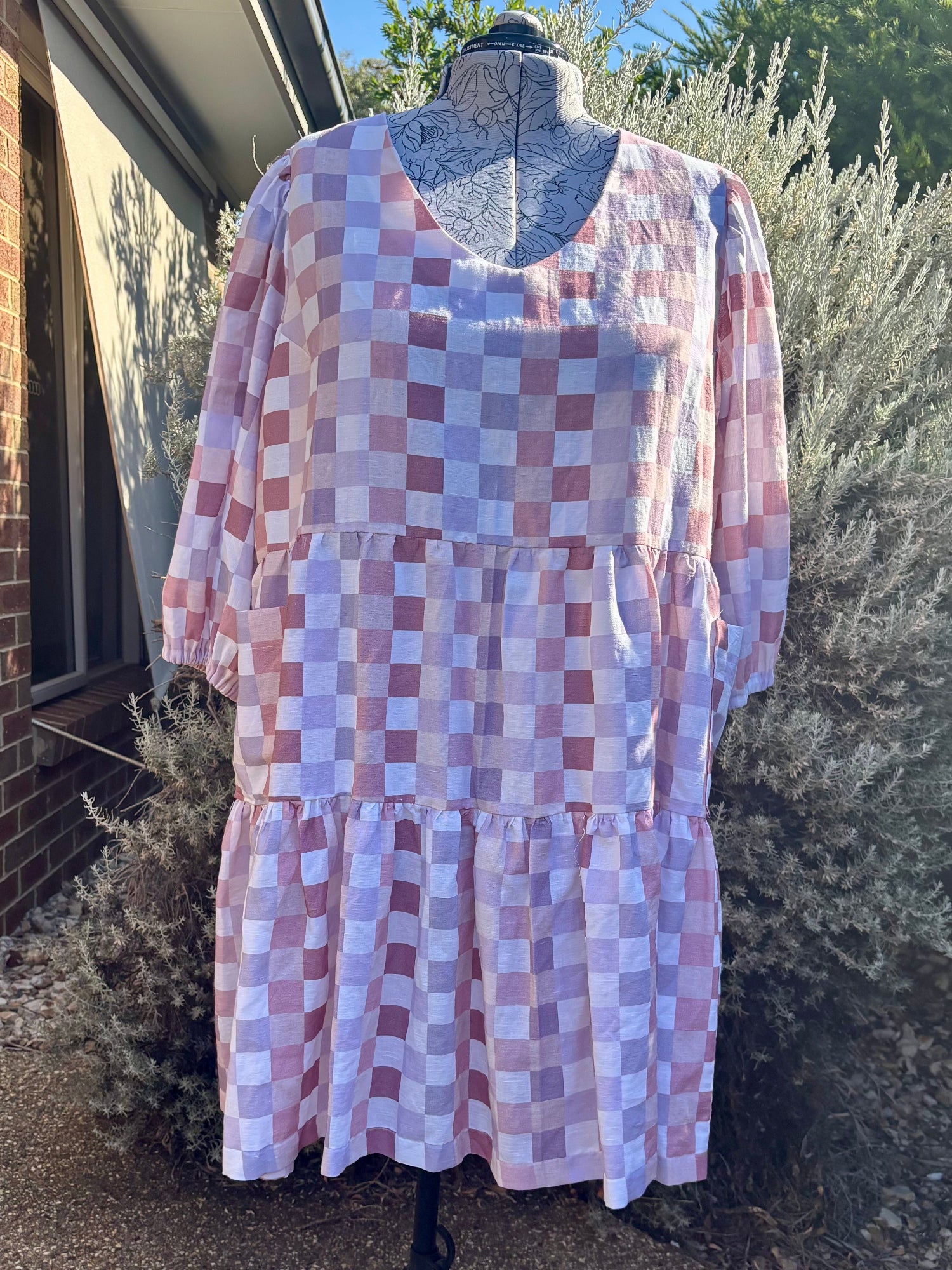 A puple checked tiered dress displayed on a mannequin, with pockets, against an outdoor background.