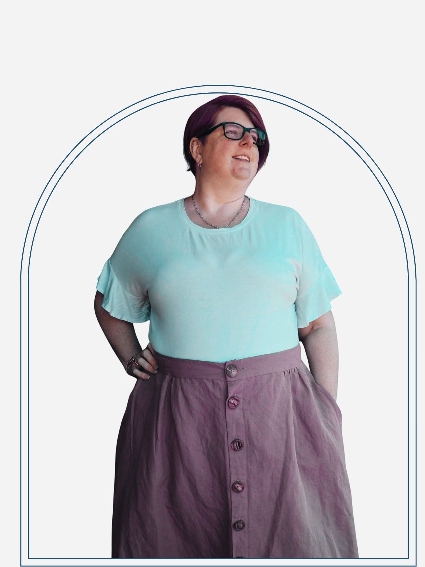 A woman standing outdoors wearing a sage green button-through skirt with natural wooden buttons, paired with a white top and glasses.